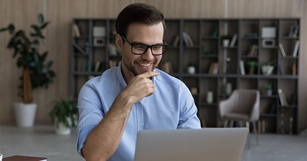 Homem de pele clara, sorrindo. ele utiliza oculos e barba. esta de camisa clara e esta vendo um notebook.